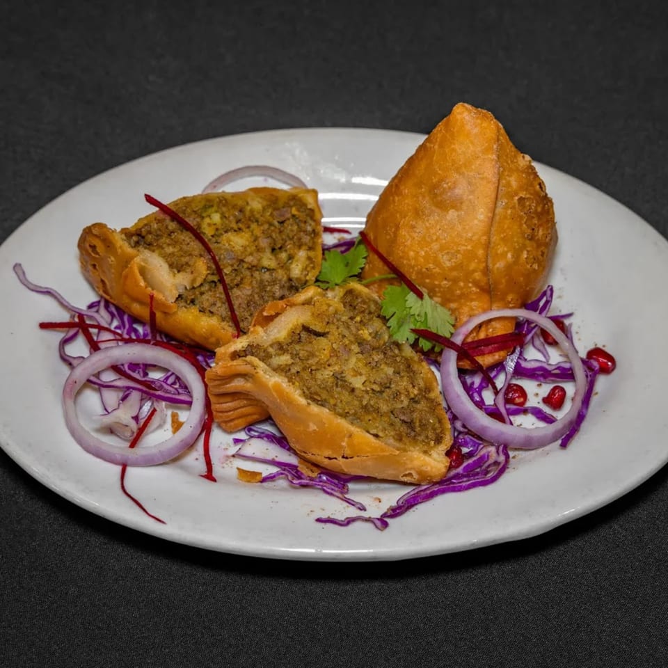 Raga Indian Cuisine's samosas, one whole and two halved, garnished with red onion, cabbage, and pomegranate seeds.