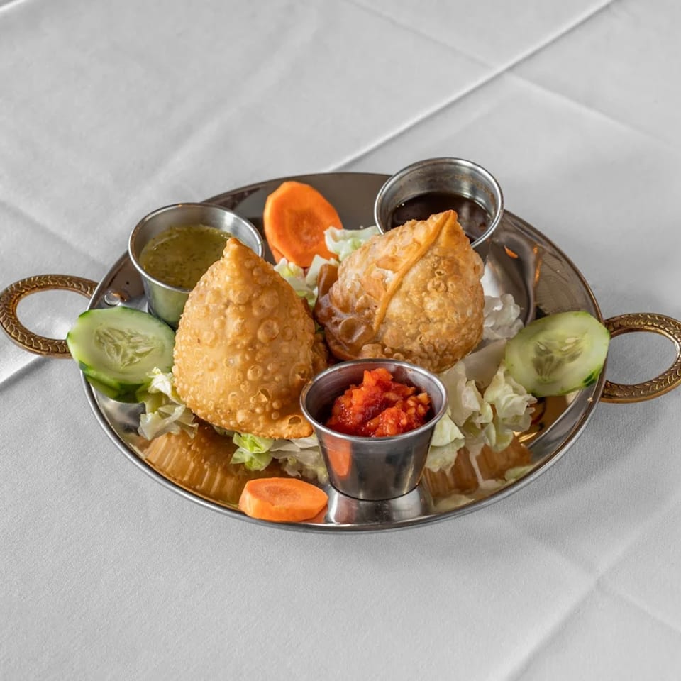 Two golden-brown samosas with three dipping sauces, lettuce, cucumber, and carrots on a silver platter.