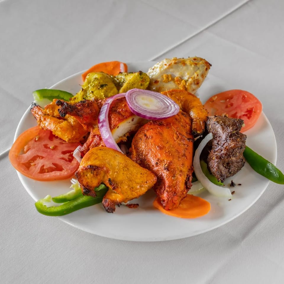 A white plate with various Indian tandoori meats, shrimp, and vegetables on a white tablecloth.