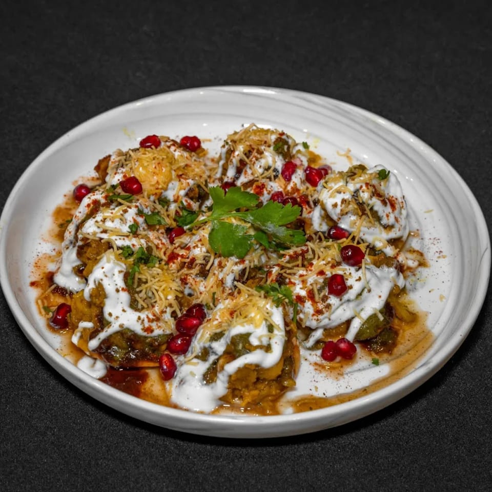 A white plate of Raga Indian Cuisine's Dahi Puri, topped with yogurt, spices, pomegranate, and cilantro.